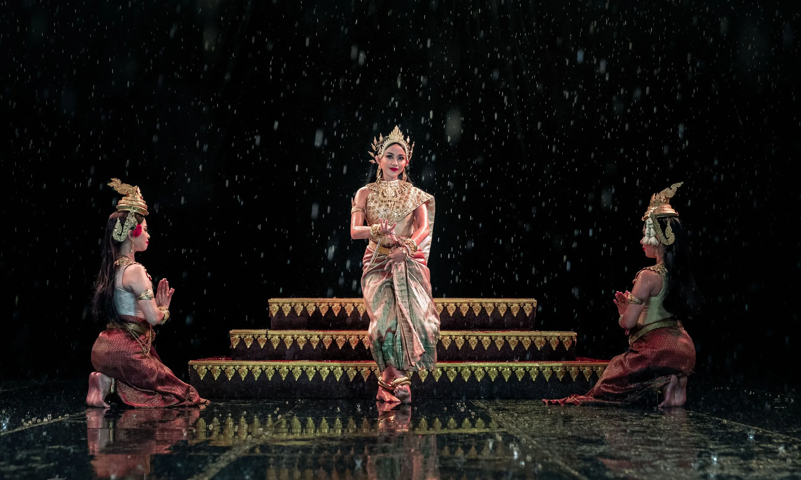 Three Cambodian classical dancers performing on a rain-soaked stage