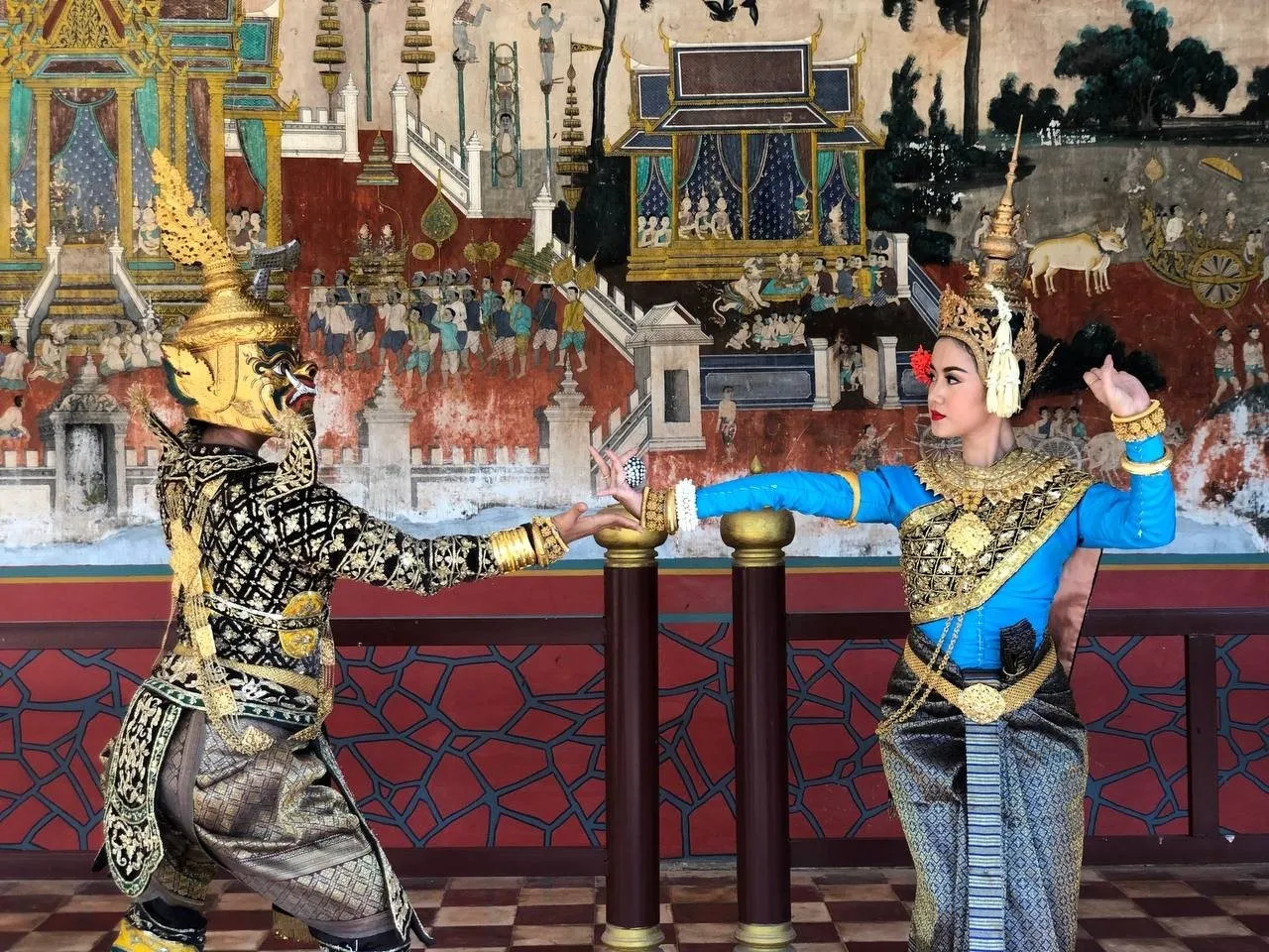 Two classical dancers, one in a masked giant role, facing each other before a painted mural